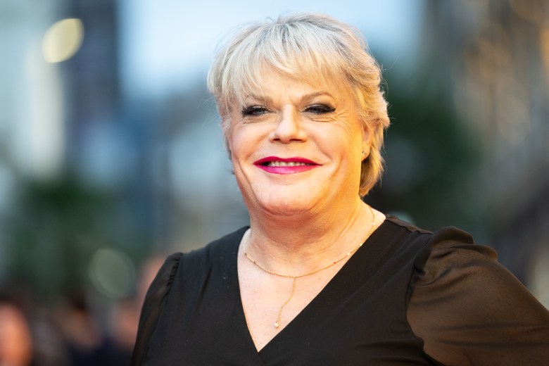 Actor Suzy Eddie Izzard smiling on a red carpet; the person has blonde hair and wears red lipstick, a black top, and a thin gold necklace. The background shows a blurred outdoor event setting.