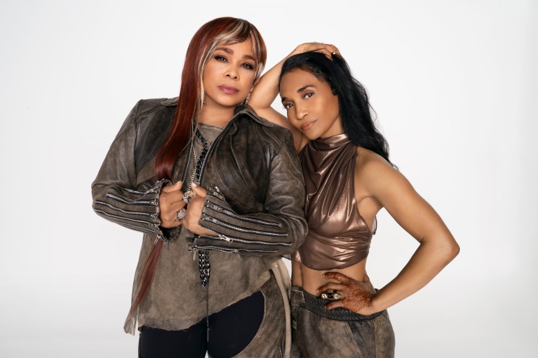 A promotional portrait of the R&B group TLC, featuring T-Boz and Chilli against a white background. On the left, T-Boz wears a distressed brown leather jacket with silver zipper details and has long reddish-blonde hair with a fringe. On the right, Chilli leans their head against T-Boz’s, wearing a metallic bronze halter top and matching trousers. Both look toward the camera with a confident, calm expression.