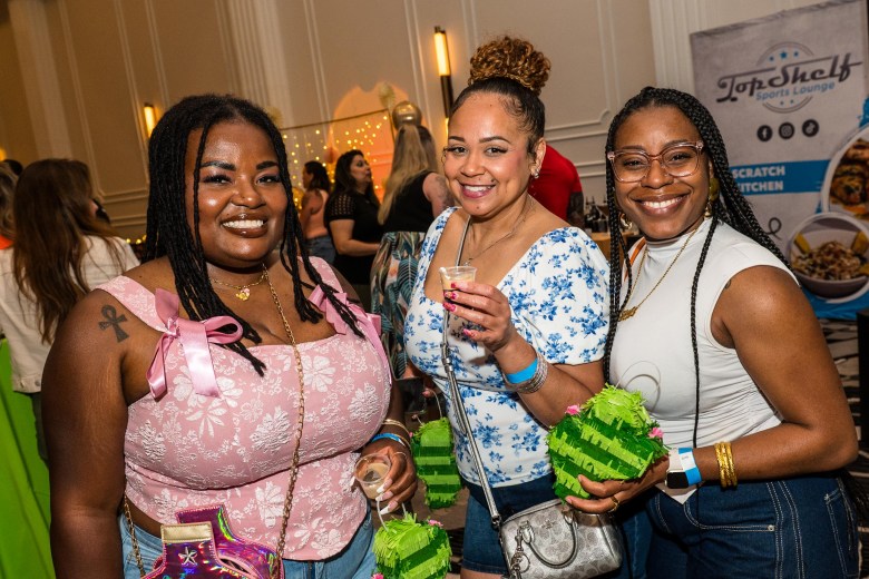 Three women smiling and holding themed green drink containers at the Taco, Tequila, and Tiki Takedown at Hotel Flor in Tampa. They are standing in a crowded event space with a "Top Shelf" branded banner in the background.