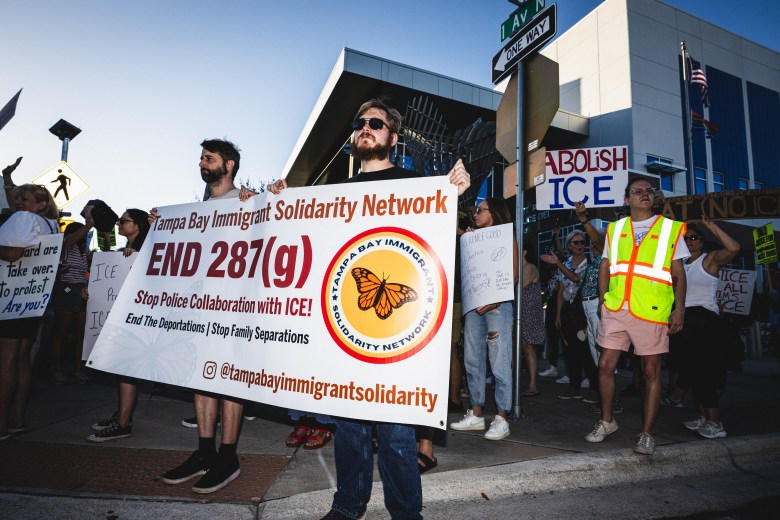 Protesters with the Tampa Bay Immigrant Solidarity Network hold a large banner that reads 'END 287(g), Stop Police Collaboration with ICE!' during an evening demonstration. The banner features a monarch butterfly logo. Nearby, another individual holds an 'Abolish ICE' sign against a backdrop of city buildings.