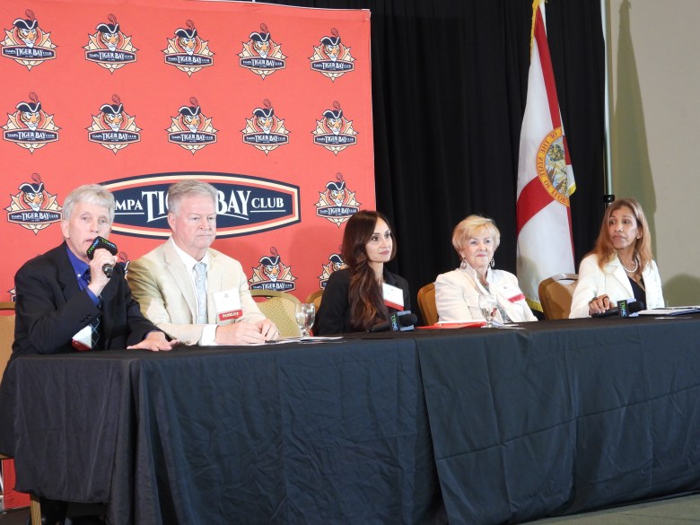 Hillsborough County School Board candidates at a Tampa Tiger Bay Club forum on March 20, 2026. Five panelists sit at a long table covered in a black cloth. From left to right: a man in a black suit speaking into a microphone, a man in a tan blazer, a woman with long dark hair in a black top, a woman with blonde hair in a white blazer, and a woman with dark hair in a white blazer. Behind them is a red backdrop featuring the Tampa Tiger Bay Club logo and a Florida state flag to the right.