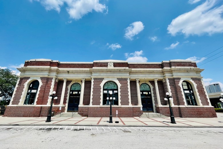 Exterior wide shot of the historic Tampa Union Station building under a bright blue sky with white clouds. The red brick structure features large arched windows, white stone accents, and a symmetrical facade with multiple dark entry doors.