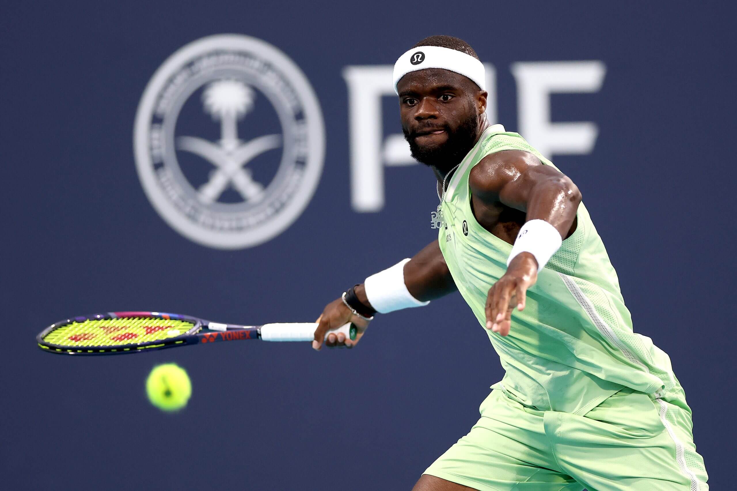 Frances Tiafoe prepares to hit a forehand wearing a green tennis outfit and a white headband.