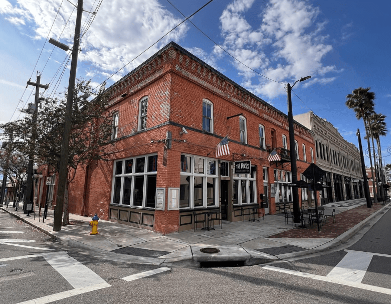 An exterior corner view of The Bricks in Ybor City under a bright blue sky with scattered clouds. The two-story historic red brick building features large windows at street level, red awnings, and American flags mounted above the entrance. The restaurant sits at a quiet intersection with white crosswalks, power lines overhead, and a row of historic buildings and palm trees extending down the sunlit street.