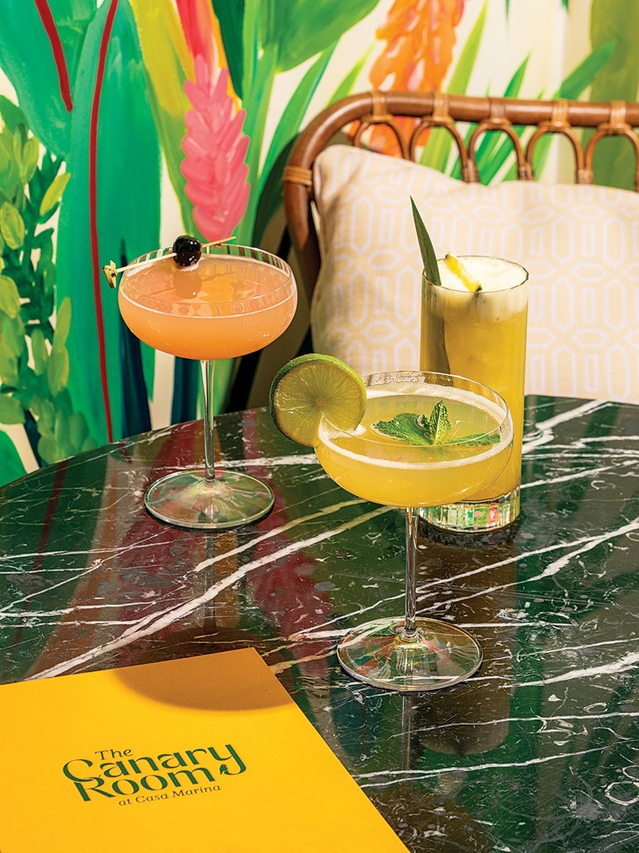 Three colorful cocktails are arranged on a dark green marble table. One cocktail is in a coupe glass garnished with a lime wheel and mint leaves, another is a peach-colored drink in a coupe glass with a cherry garnish, and the third is a tall yellow drink garnished with a pineapple leaf and a pineapple wedge. A yellow menu with 