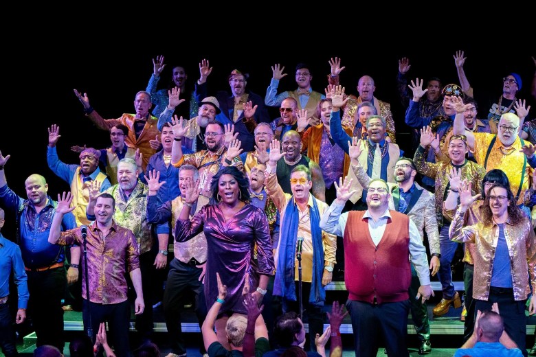 The Gay Men's Chorus of Tampa Bay performing on stage during the 'Say My Name' cabaret at the Straz Center. A diverse group of singers is spread across the stage, many wearing shimmering gold and patterned jackets. A person in a metallic purple dress stands at the center-front, and all performers have their hands raised in an expressive, synchronized gesture against a dark background.