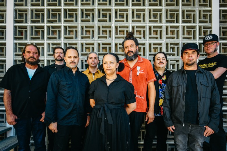 The Sh-Booms band members posing together in front of a white geometric breeze block wall. The eight individuals are dressed in stylish casual and professional attire, featuring a mix of black button-downs, a gold shirt, and a central figure in a black wrap dress.