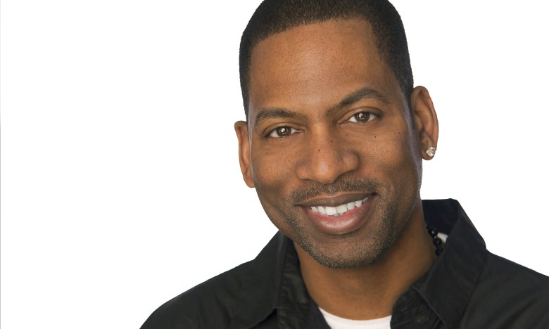 A close-up professional headshot of comedian Tony Rock against a white background. The person is smiling and wearing a black button-down shirt over a white undershirt. A small diamond stud earring is visible in their left ear, and they have a neat goatee and short-cropped hair.
