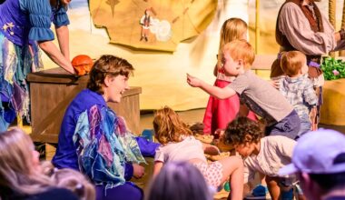 A young blonde boy shows an object in his hand to a performer in a blue and purple costume, surrounded by an audience