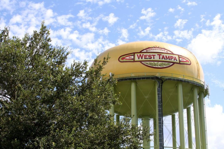 Low-angle view of the historic West Tampa water tower, painted yellow with a cigar-label style logo, set against a bright blue sky with light clouds and green tree foliage in the foreground.