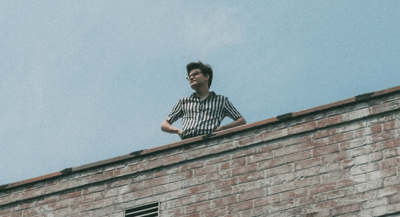 Musician Zack Keim wearing a black and white striped short-sleeve shirt, leaning against a weathered brick wall under a clear blue sky. The image has a grainy, film-like texture.
