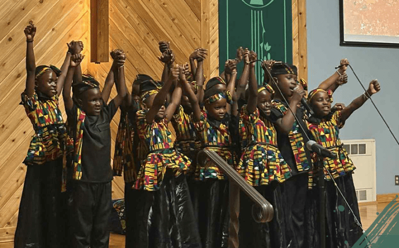 Members of the African Children's Choir performing on a wooden stage. The children are dressed in vibrant, matching outfits featuring yellow, red, and blue geometric patterns. They are captured mid-performance with their arms raised high and hands held, some singing into a microphone against a backdrop of light wood paneling