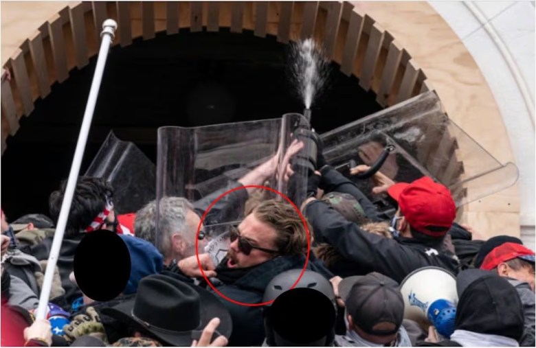 A man with brown hair and sunglasses, circled in red, amidst a crowd of individuals clashing with police and using riot shields at a stone archway during the January 6 Capitol riot.