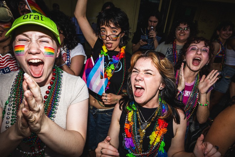 An energetic crowd of young people wearing rainbow face paint and colorful beaded necklaces , with some waving trans pride flags and cheering wildly at Tampa Pride 2025.