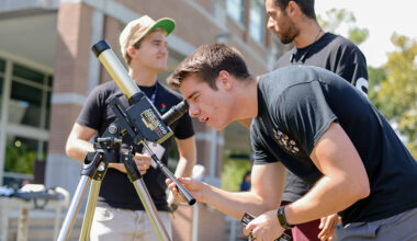 Man wearing black shirt bends down to look through telescope. There are two other men standing behind him talking.