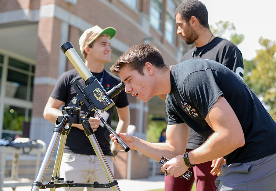 Man wearing black shirt bends down to look through telescope. There are two other men standing behind him talking.