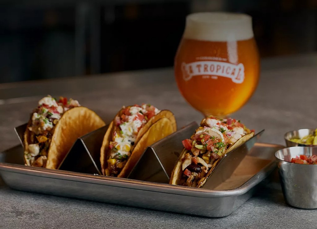an orange craft beer in a class besides a row of three tacos with toppings