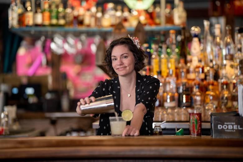 A person with short curly hair and a flower accessory pouring a drink from a metal shaker into a glass at the Casa Tina bar in Dunedin.