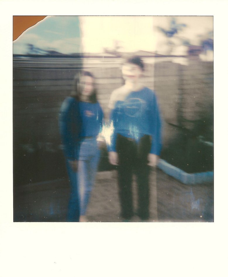 A blurry, overexposed Polaroid photo of the two members of the band Catnap; both individuals wear blue long-sleeve shirts and stand outdoors in a backyard setting with a wooden fence and palm trees in the background.