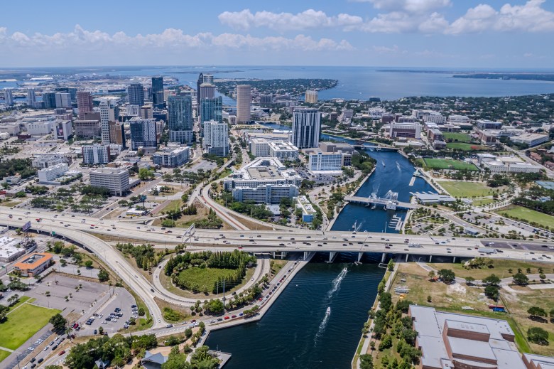Aerial view of the Downtown Tampa skyline and the Hillsborough River on a clear, sunny day. The image shows high-rise office buildings, the University of Tampa, and a major highway interchange with cars crossing the river into the urban center.