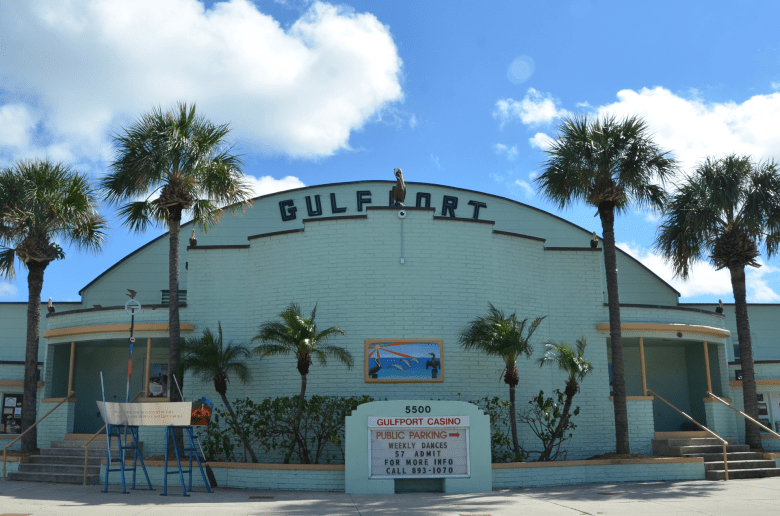 The historic Gulfport Casino Ballroom in St. Petersburg, Florida; a lime-green Art Deco style building with palm trees and a sign board, situated on the waterfront of Boca Ciega Bay.
