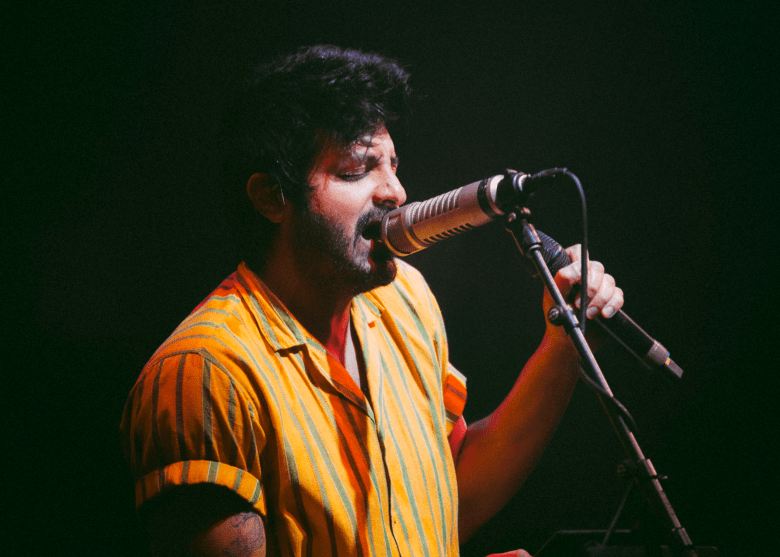 A live concert photograph of Sameer Gadhia, lead singer of Young The Giant, performing on stage. They have dark curly hair and a beard, wearing an orange and yellow vertical-striped short-sleeve button-down shirt. They are leaning into a vintage-style silver microphone, eyes closed in a moment of intense vocal performance against a dark, moody background.