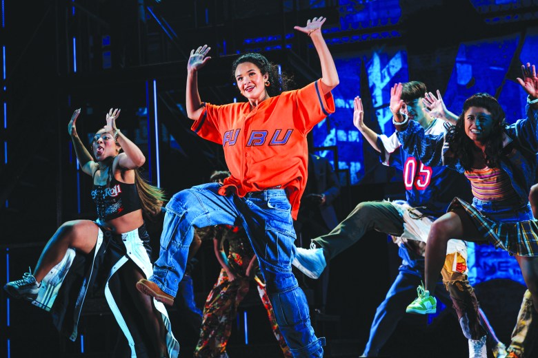 A vibrant group of dancers performing a hip-hop routine on stage, led by Maya Drake in a bright orange FUBU jersey for 'Hell's Kitchen'