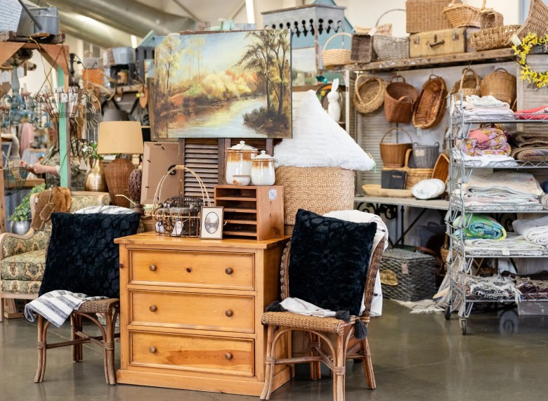 A curated display of vintage furniture and home decor, including a wooden dresser, wicker chairs, and various woven baskets at an indoor antique market.