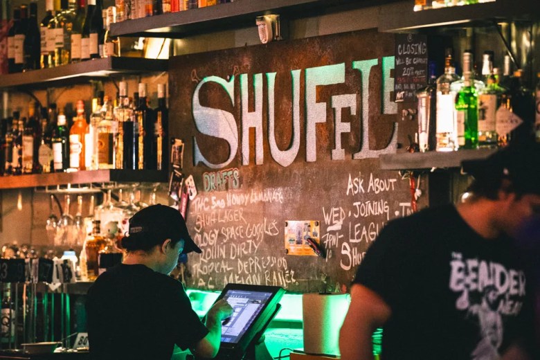 Bartenders working behind the counter at Shuffle in Tampa, featuring a large chalkboard menu with craft beer drafts like 'Tropical Depression' and 'Rollin Dirty'.