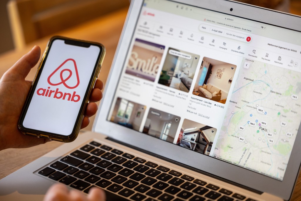 Female hand holding a smartphone displaying the Airbnb logo next to a laptop showing the Airbnb website with available lodgings in Paris.