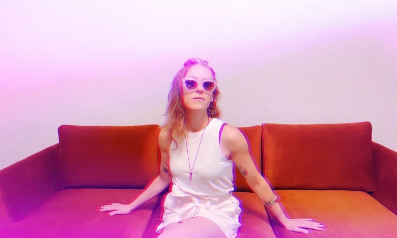 Songwriter Katie Costello sits on a plush burnt-orange sofa against a plain wall. The person wears a white sleeveless top, a thin necklace, and pink-rimmed sunglasses. The image is bathed in a soft pink and purple neon glow, creating a dreamlike, cinematic atmosphere.