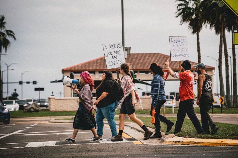 A line of activists walking through a crosswalk outside MacDill Air Force Base, with one person speaking into a megaphone and others carrying signs that read 'ORDERS ARE NOT AN EXCUSE' and 'End U.S. Imperialism'."