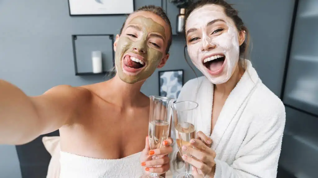 Photo of a happy cute amazing women at home in towel and bathrobe after spa procedure taking care of their skin drinking champagne take selfie by camera.