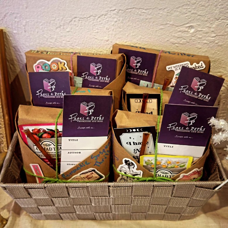 Several book bundles from Pages and Perks bookstore wrapped in brown kraft paper and tied with green and pink twine. Each bundle includes a purple bookmark with the shop's logo, a 'Blind Date with a Book' label with genre descriptions like 'Romantic Suspense' and 'Thrill,' and a packet of 'Mad Tea' herbal tea."