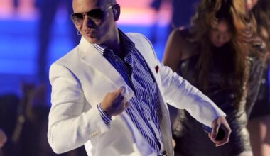 photo of singer Pitbull wearing a button-down shirt and white blazer jacket with a backup dancer behind him onstage