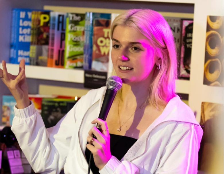 A person performing stand-up comedy at a microphone in a bookstore, gesturing with their hand in front of a bookshelf featuring titles like 'If You Love Let Kill' and 'The Great Good.'