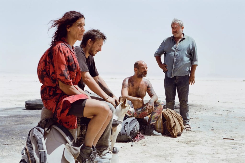 still from the movie "Sirât" showing four disheveled people sitting on backpacks on the beach