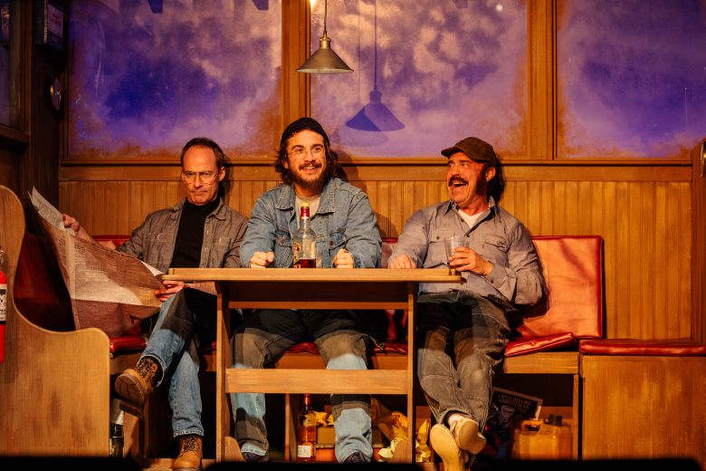 Production still from 'The Shark Is Broken' at Jobsite Theater. From left to right: Christopher Marshall (as Roy Scheider) reads a newspaper, Cody Farkas (as Richard Dreyfuss) sits at the center of a wooden table with a bottle, and Paul Potenza (as Robert Shaw) laughs while holding a drink. They are seated in the wood-paneled galley of a boat with blue-tinted windows in the background, captured during a March 2026 rehearsal in Tampa.