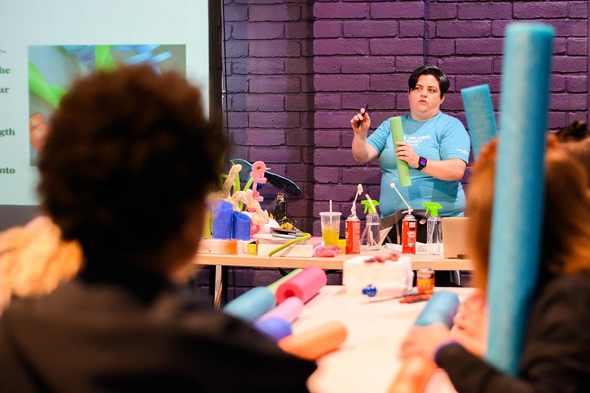 Woman wearing teal t shirt stands behind a table with various crafting supplies and holds up a green pool noodle and pen.