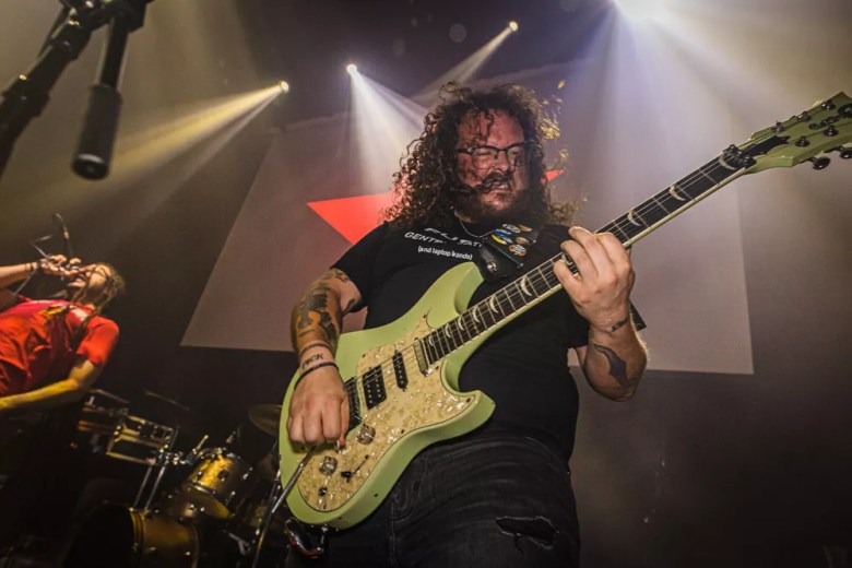 Musician Justino Lee Walker of Anthill Cinema performing live on stage. The person has curly dark hair and glasses, wearing a black t-shirt while playing a light green electric guitar. In the background, another performer in a red shirt is visible under bright stage spotlights and a large red star graphic.