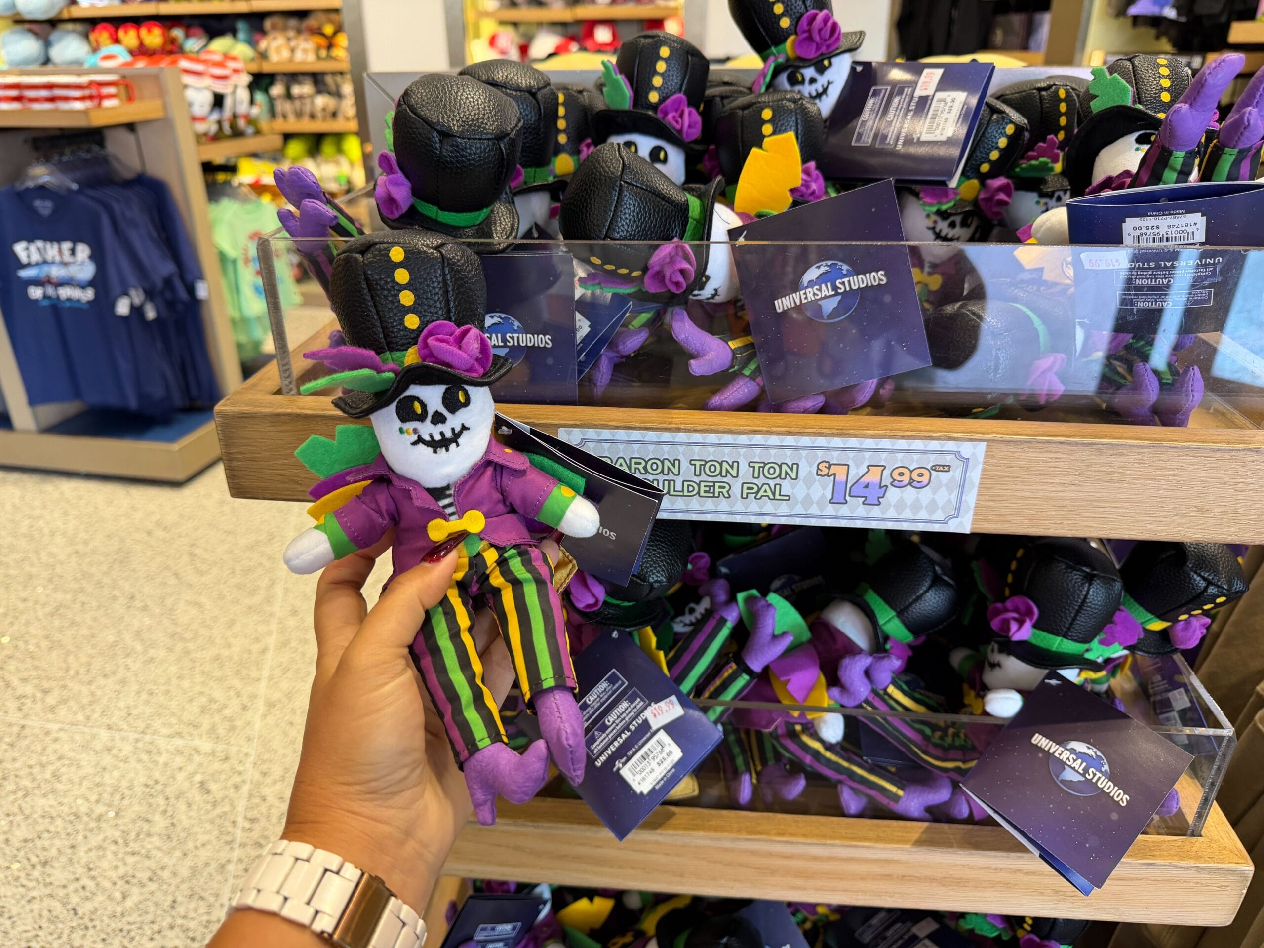 This shoulder plush is designed to look like the Baron, another mascot of the Mardi Gras season at Universal Studios. He has a cheerful skull face and is wearing a black top hat, a purple coat, and multicolored striped pants.
