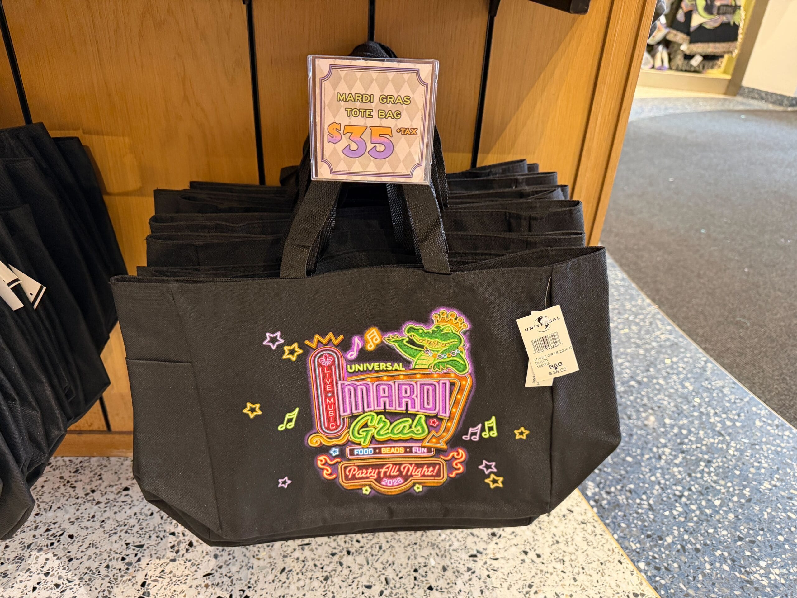 This black tote bag has a neon sign-themed design on the front featuring King Gator and the 2026 Universal Mardi Gras logo.