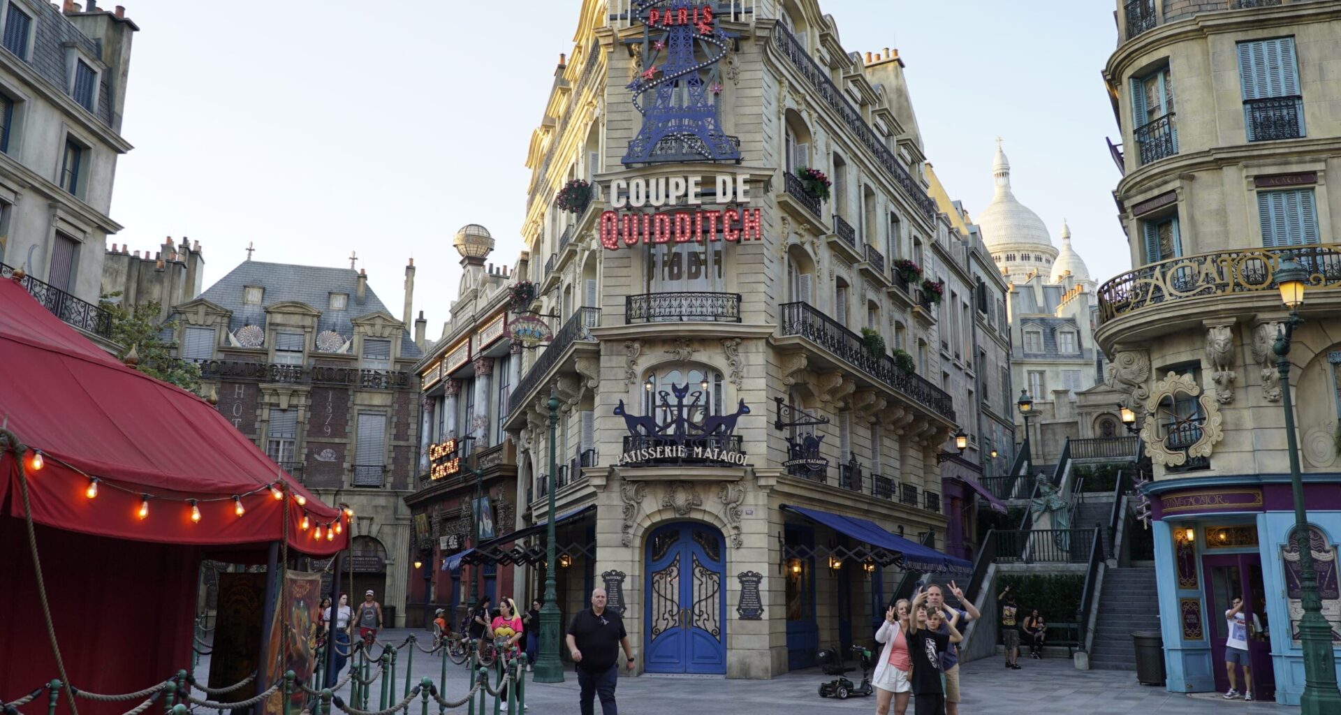 Guests stroll and snap photos in a city square with ornate fa&ccedil;ades, a Quidditch sign, and Daily Recap display&mdash;no castle in sight.