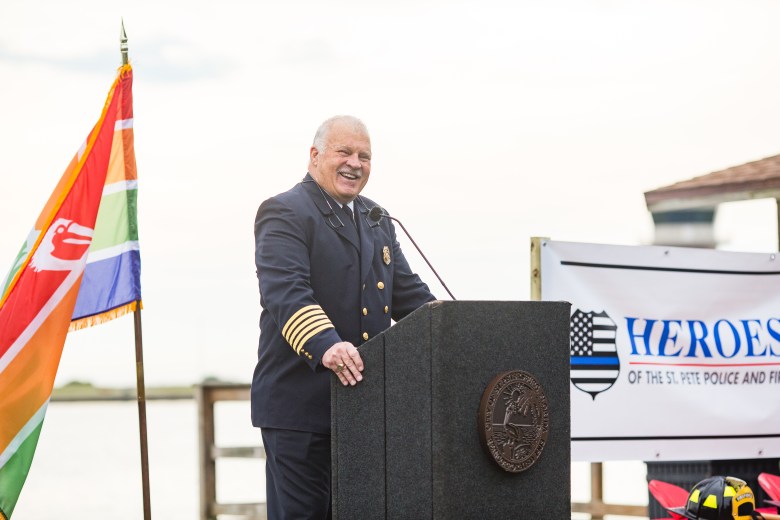 Former St. Petersburg Fire Chief Jim Large smiling at a podium during the Heroes of St. Pete Fire Memorial groundbreaking ceremony. He is in his full dark blue dress uniform with gold captain's bars on the sleeves, standing beside a colorful City of St. Petersburg flag with the waterfront in the background.