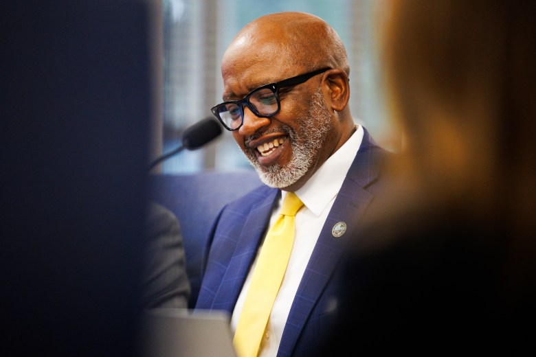 St. Petersburg Mayor Ken Welch smiling and wearing glasses and a yellow tie while seated at a committee table. The "We Are St. Pete" branding is partially visible in the blurred foreground, with a professional city hall setting in the background.