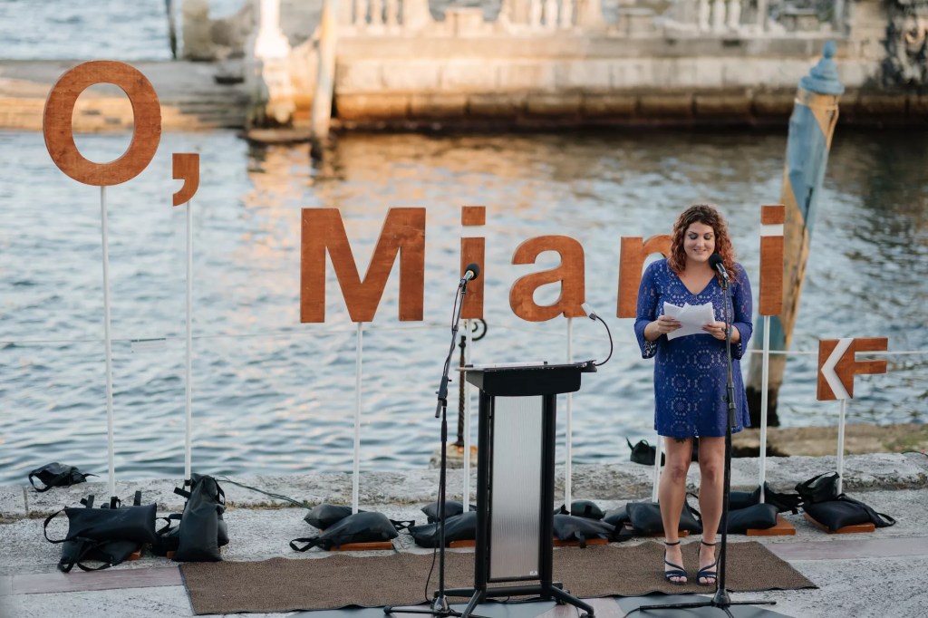 a person reads a poem in front of the water and a backdrop that reads, "O, Miami"