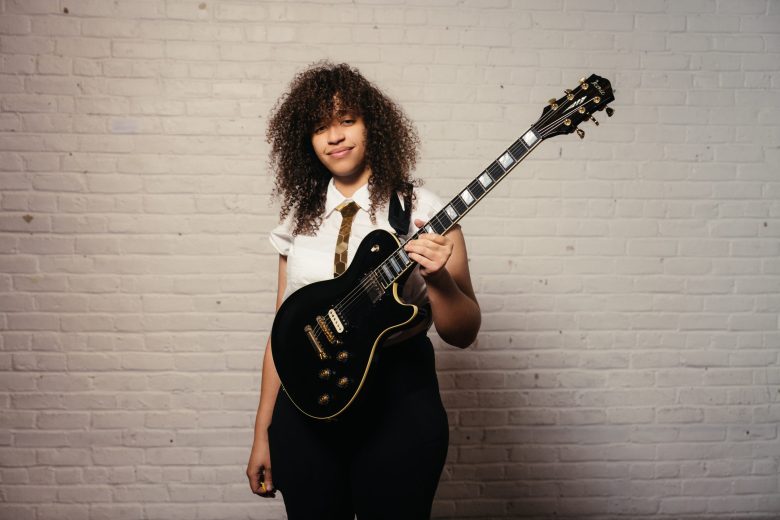 Musician Jackie Venson stands in front of a white brick wall, holding a black electric guitar. She is wearing a white short-sleeved button-down shirt with a tan tie and dark trousers, looking at the camera with a confident expression. Her signature voluminous dark hair is a focal point of the portrait.