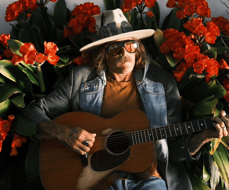 Singer-songwriter Donavon Frankenreiter sits playing an acoustic guitar in front of a dense wall of bright orange flowers. He is wearing a wide-brimmed cream hat with a decorative band, tinted sunglasses, a denim jacket over an orange shirt, and has a prominent handlebar mustache.