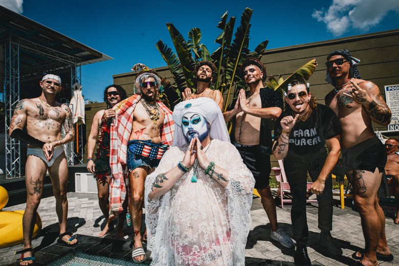 A group of contestants poses poolside at the Wet Spot during the Hunky Jesus contest. In the center, a person in "Foxy Mary" drag wears a white lace veil and dress with blue-and-white face makeup, holding their hands in prayer. They are flanked by several "Hunky Jesus" contestants in swimwear, some shirtless and showing tattoos, under a bright blue sky with palm trees in the background.