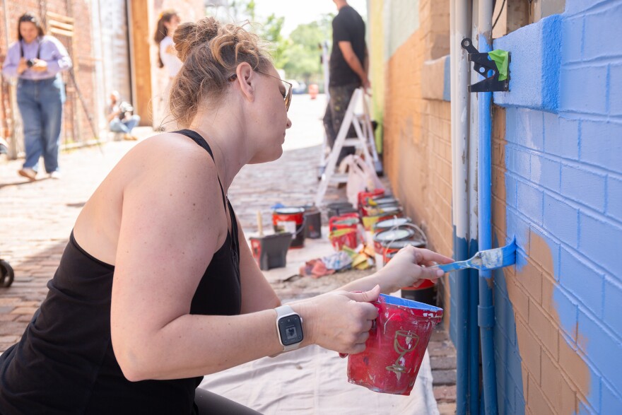 A woman with a black tank top and blonde bun and sunglasses kneels down next to a brick wall to paint a large swathe of blue. Behind her and slightly out of focus are rows of open paint cans.
